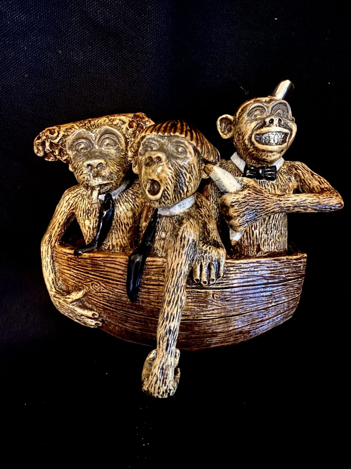 Titled A Barrel of Fun!, these three whimsical bronze monkey figures in a small boat depict see no evil, hear no evil, speak no evil. Adorned with bow ties, they are crafted from textured material for an antique appearance set against a dark background.