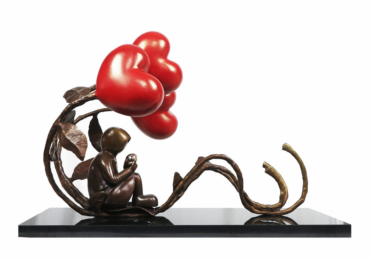All The Love In The World is a bronze sculpture by Mackenzie Thorpe featuring a child seated, gazing at a large red heart-shaped flower on a curving vine, set on a black base against white—showcased in the Lake Tahoe art district.