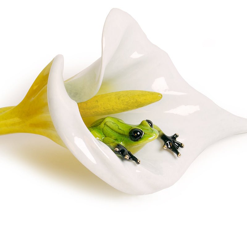 A small green frog with black-tipped toes nestles inside the Calla Lily sculpture by Tim Cotterill, blending naturally with the flowers smooth petals against a pristine white background.