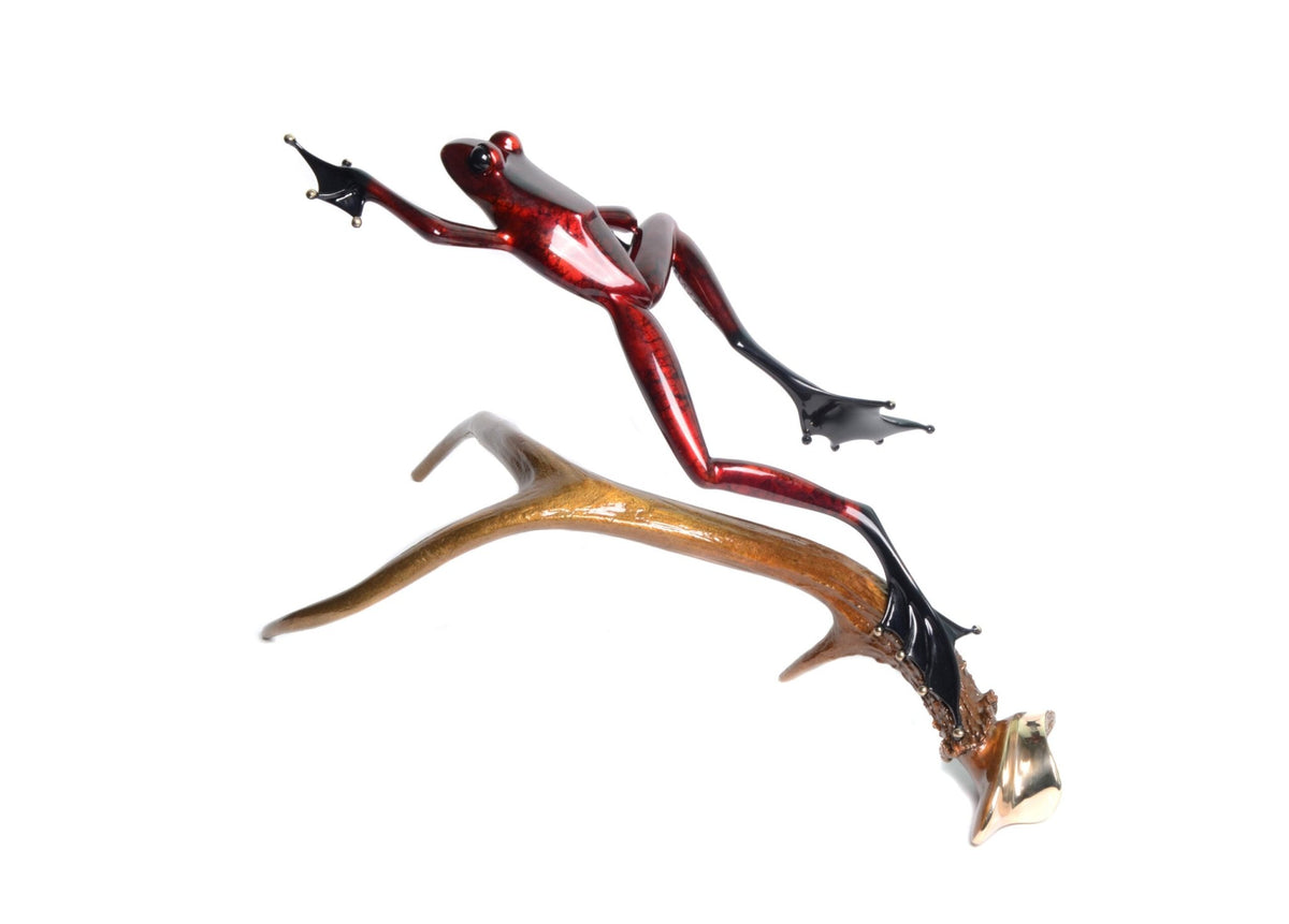 The Stags Leap is a glossy red Frogman Bronze sculpture with elongated limbs and black webbed feet, artfully poised as if leaping on a smooth, curved branch-like structure. It stands out beautifully against the white background, emphasizing its intricate details and vibrant colors.