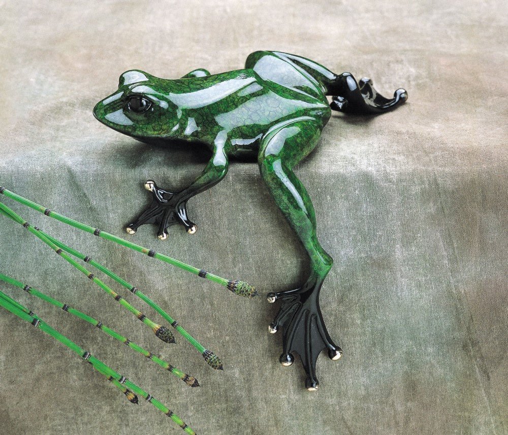 A shiny Fat Boy Bronze sculpture of a frog with black feet sits on a textured surface. Surrounding it are elongated green reeds with black accents, adding natural charm. The frogs emerald green patina and polished surface reflect light beautifully.