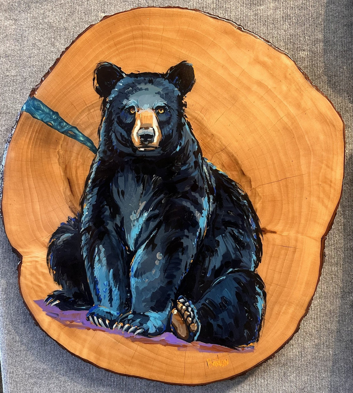 This Steve Barton original, Baxter, is a vibrant painting of a black bear on a tree slice, featuring blue and black hues with realistic expression. The woods natural rings and textures provide a rustic backdrop to this captivating piece of art.
