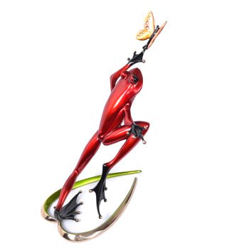 The Be Mine sculpture features a sleek red metallic frog with long limbs and black feet, reminiscent of Frogman art. Balancing on two curved leaf-like structures, the frog reaches for a delicate butterfly on its hand. A plain white background accentuates its elegance.