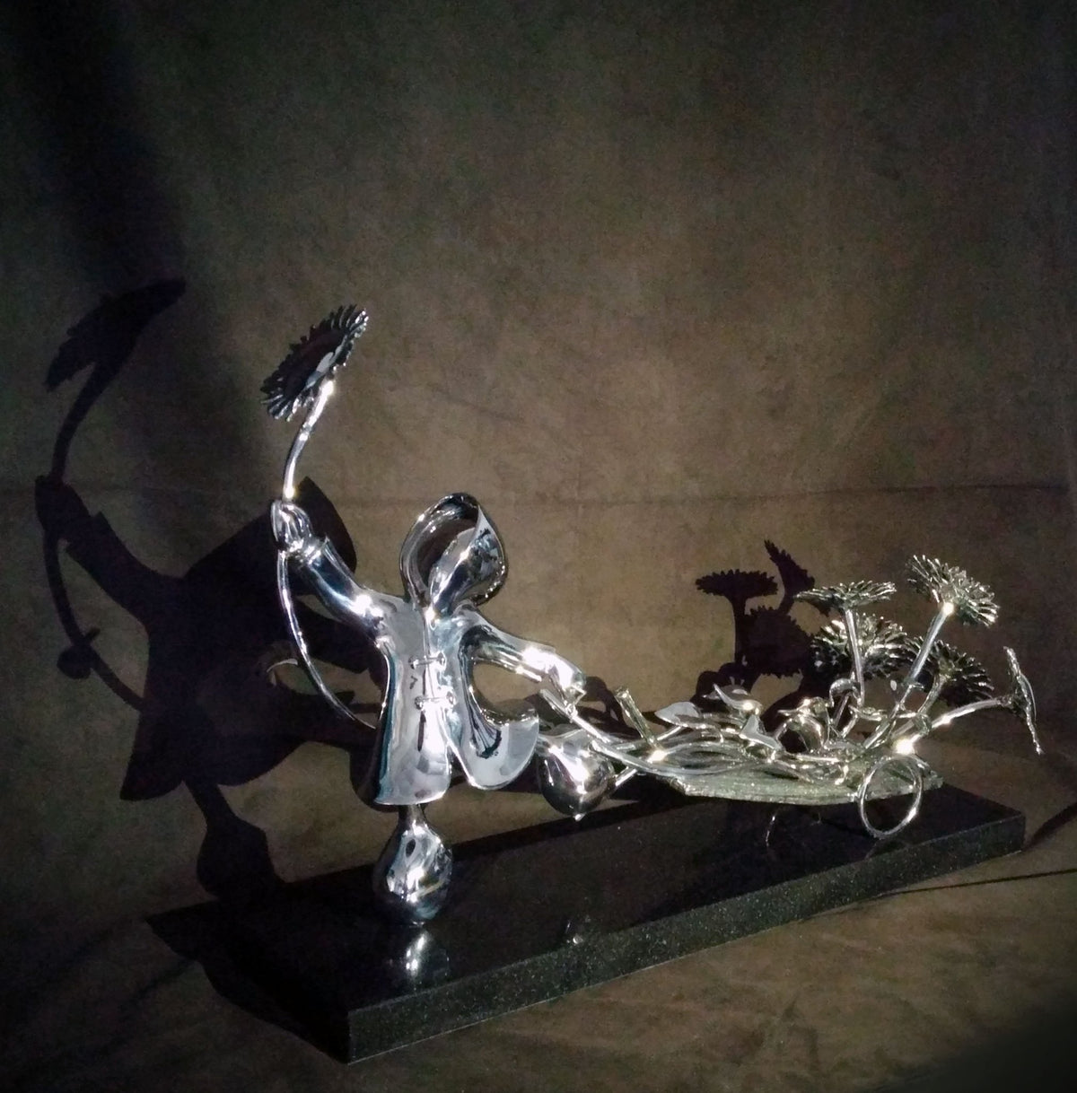The limited edition Bringer of Sunshine-Stainless by Mackenzie Thorpe features a whimsical figure holding a sunflower, with more sunflowers on its base. The artworks shadow on a plain background enhances its playful essence.