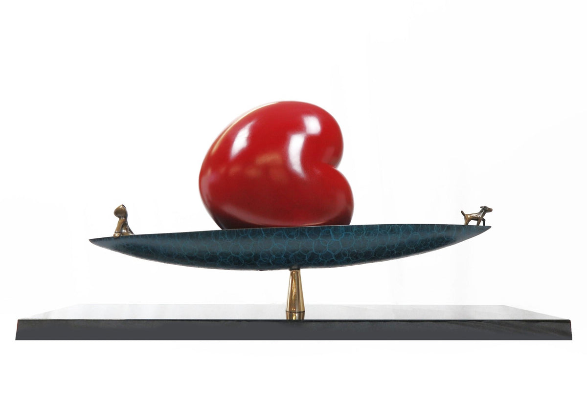 The Calm Waters sculpture by Mackenzie Thorpe features a large, glossy red heart on a teal and bronze dish with small dog and cat figurines at each end, creating harmony. Displayed at Marcus Ashley Gallery, it rests on a sleek black rectangular base.