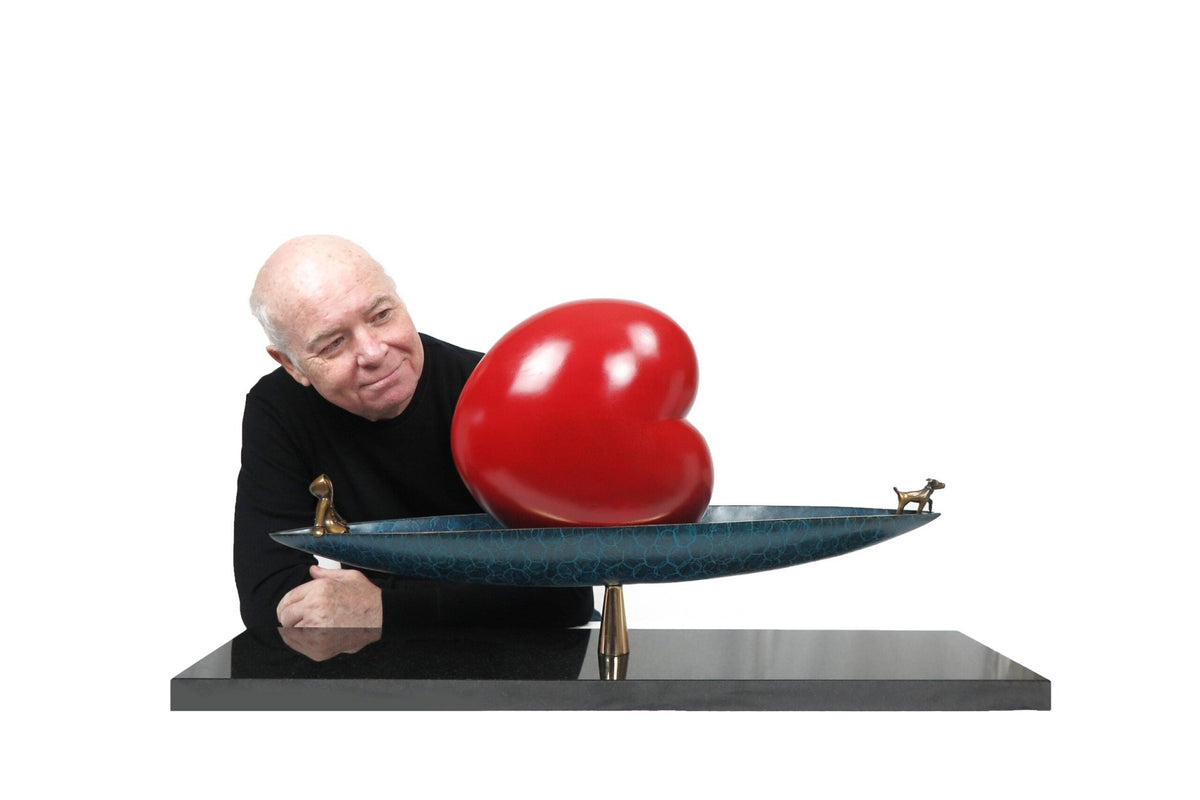 A person in a black shirt admires the Calm Waters sculpture, an evocative piece by Mackenzie Thorpe featuring a glossy red heart on a teal boat-shaped base adorned with tiny animal figurines. Its displayed at Marcus Ashley Gallery against a white backdrop.