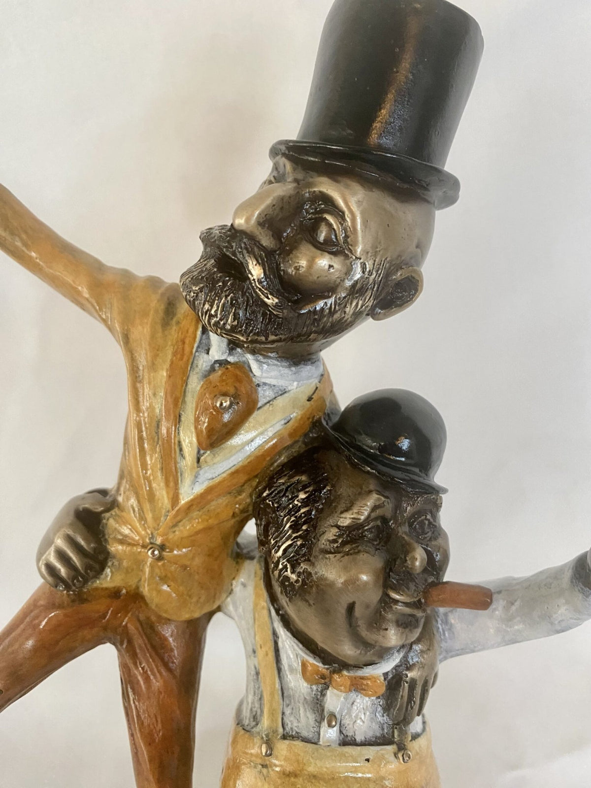 The Cheers bronze sculpture by Paul Lotz depicts two jovial men with exaggerated features: one in a top hat and beard, the other in a bowler hat and cigar. Dressed in vintage attire with bow ties, they strike a lively pose full of whimsy.