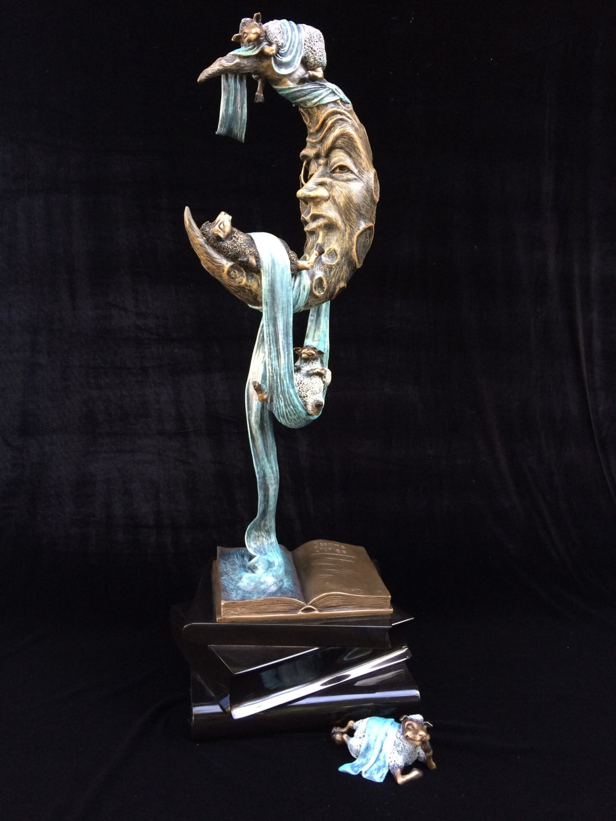 Counting Sheep is a whimsical bronze sculpture combining abstract and realistic elements, featuring a crescent moon with detailed faces, flowing blue fabric, small human figures, a polished black base with an open book and another figure nearby, evoking a bedtime ritual scene.