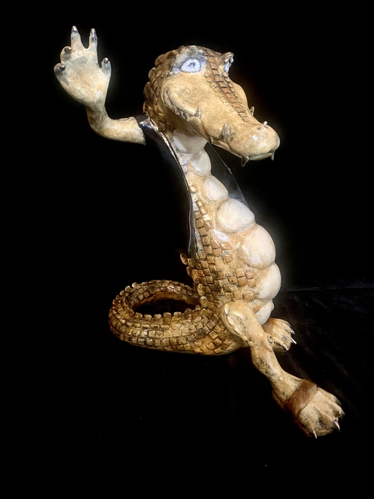 Paul Lotzs whimsical bronze sculpture, Crocodile Rock, showcases a cartoonish crocodile with an exaggerated expression. It sits upright on a plain black background, waving one arm, with a textured brown surface and big playful eyes.