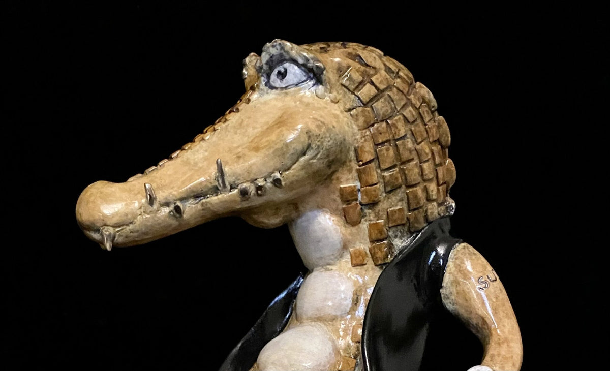 The Crocodile Rock ceramic sculpture showcases a smiling alligator with a textured, checkered body and head, glossy black vest, and a prominent snout with visible teeth. Its intricate details stand out against the black background.