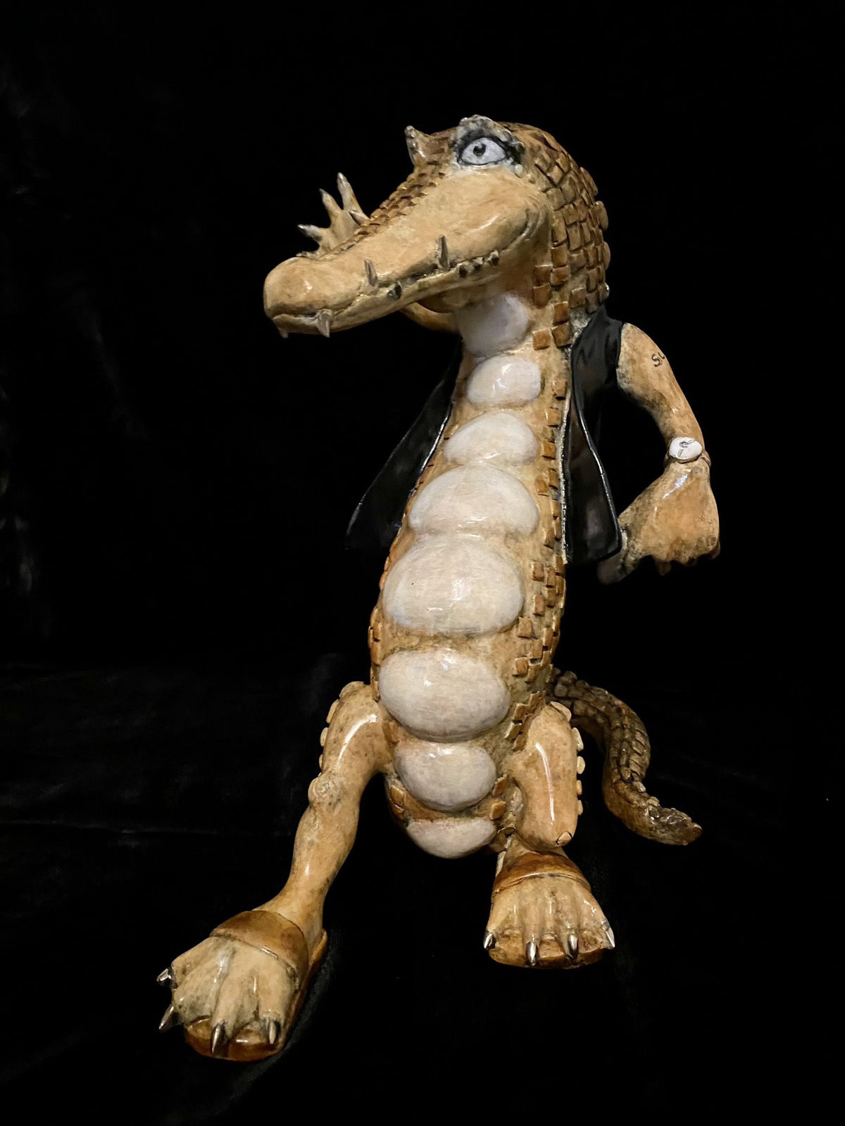 The Crocodile Rock ceramic figure, inspired by Paul Lotz, stands upright with textured scales and a prominent belly. It wears a black vest and white wristwatch against a black background, while its large eyes add charm to the whimsical design.