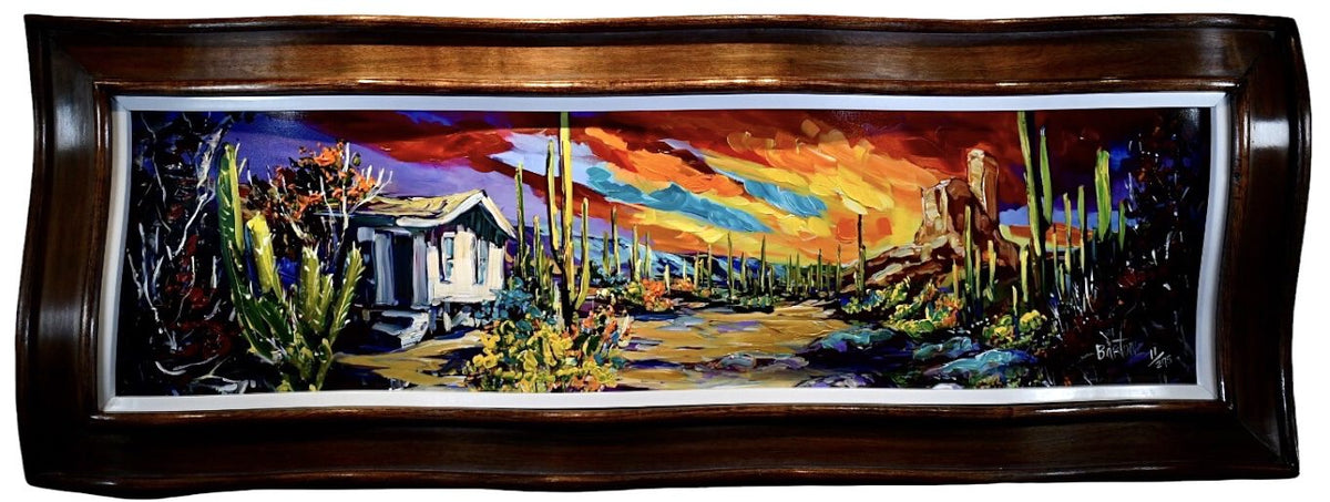 The Desert Sky by Steve Barton, enclosed in a dark wooden frame, showcases a Southwest desert scene with towering cacti, a quaint building, and a vivid sunset of red, orange, and purple hues. Its bold texture vividly captures the dramatic essence typical of Bartons work.