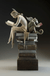 The bronze sculpture Ex Libris by Michael Parkes features a winged nude figure and a winged cat sitting back-to-back on books. The human reads an open book, and the cat holds another, symbolizing themes of curiosity and knowledge.