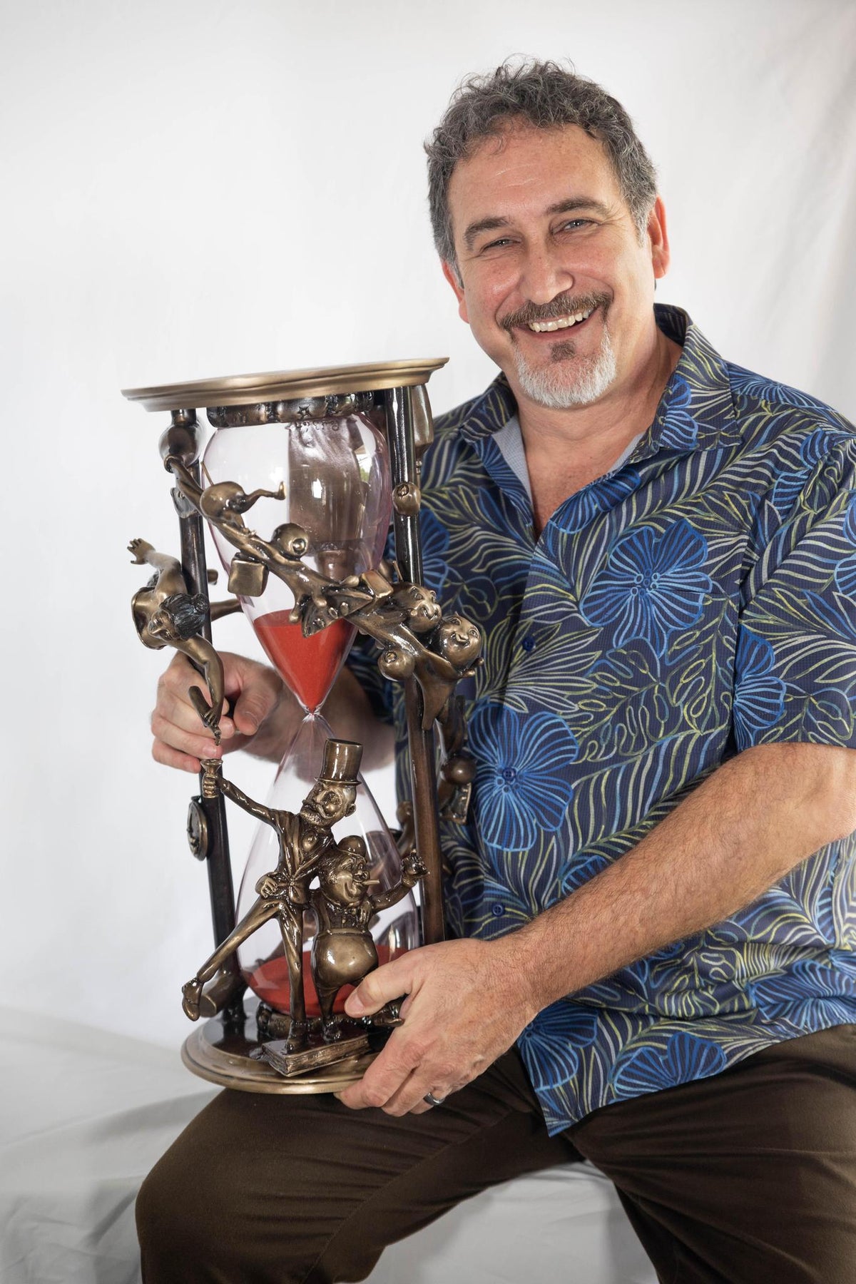 A smiling gray-haired man with a beard wears a blue patterned shirt while holding Happy Hour, an ornate hourglass with bronze figures climbing its frame and red sand inside, set against a plain white background.