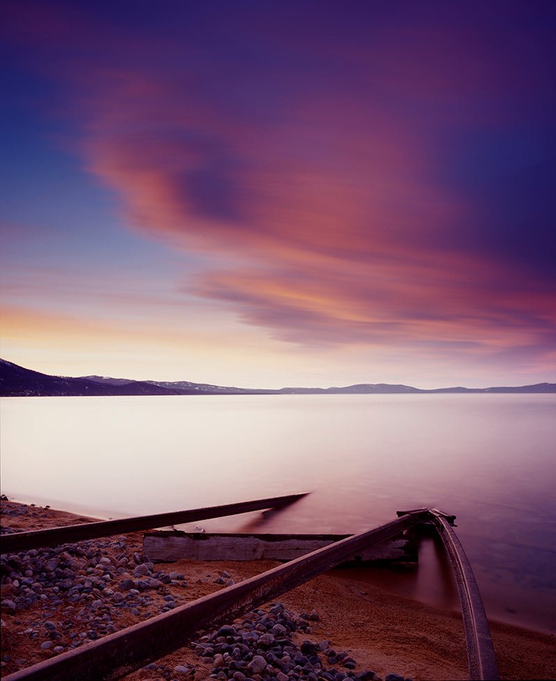 Captured in the Historic Tracks and Sunset photo, vibrant purple and orange clouds stretch across the Lake Tahoe sky at dusk. Calm waters reflect this scene, bordered by a rocky shore and old wooden beams reminiscent of historic tracks extending into the lake.