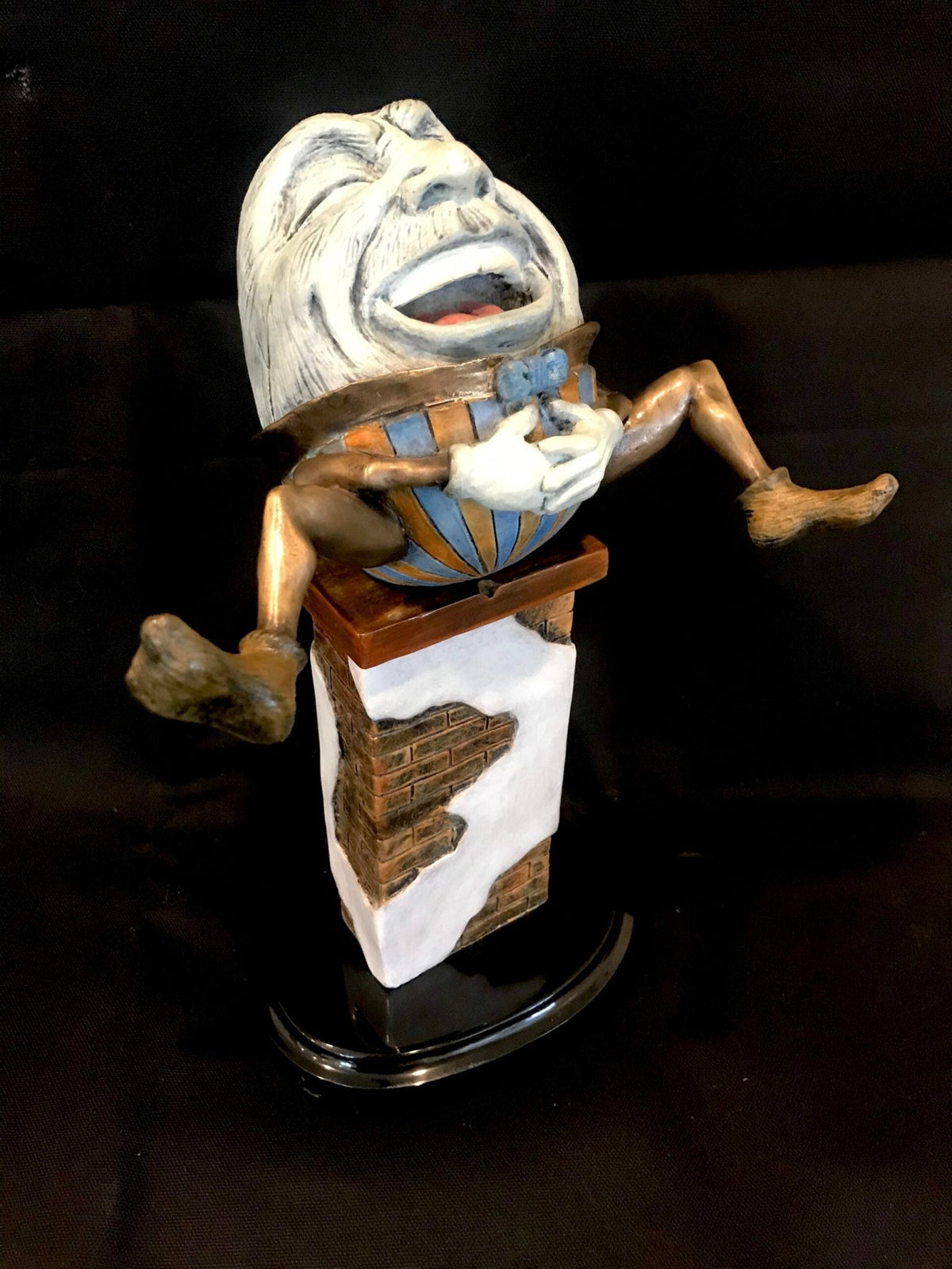 The Humpty w/ bronze wall base features a lively depiction of Humpty Dumpty joyfully laughing atop a brick wall. His expressive face and outstretched limbs capture the whimsy of the beloved nursery rhyme, enhanced by intricate details against a dark backdrop.