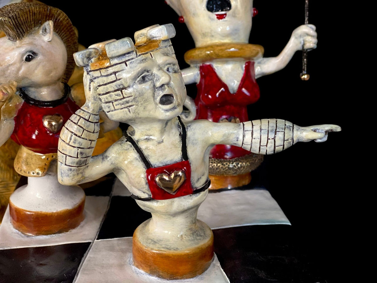 The White Rabbit is a ceramic chess piece shaped like a castle, with a human face, one arm raised to its ear and the other pointing. Dressed in red with a gold heart, it evokes Alices whimsical world amid quirky chess characters.