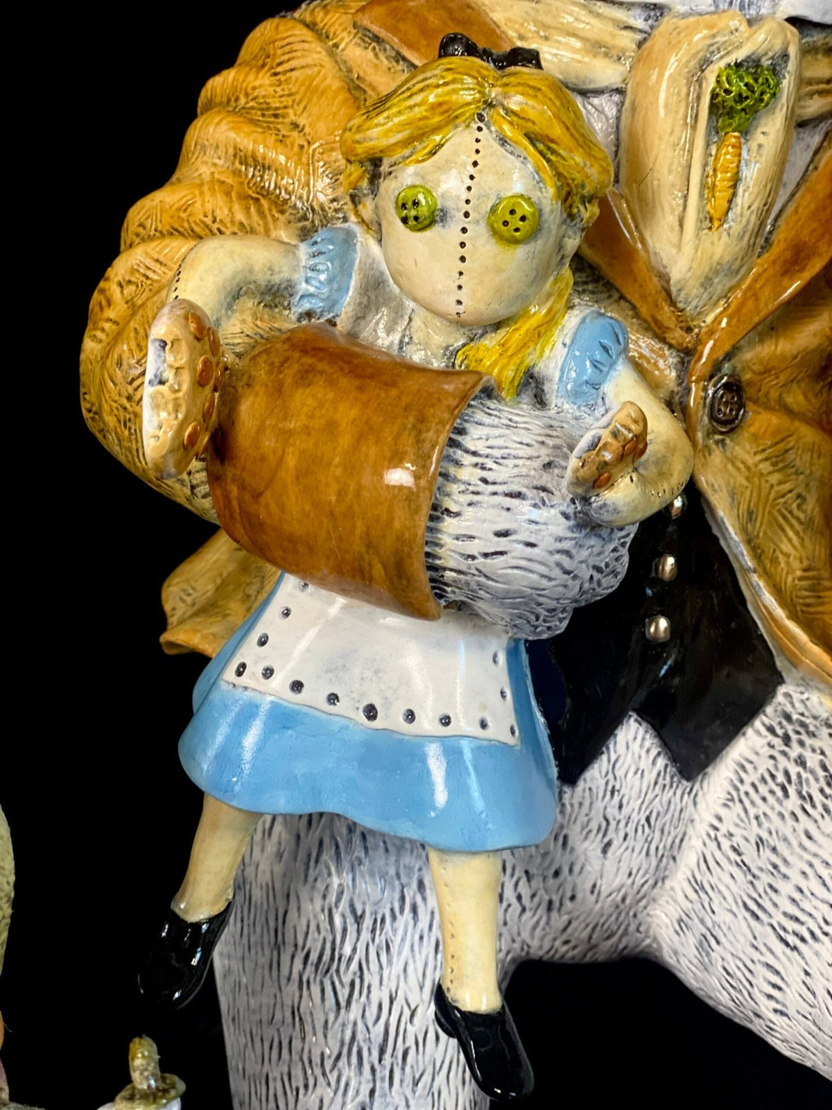 The White Rabbit features a ceramic Alice figure with button eyes, blue dress, white apron, and yellow hair, held by a larger figure in a brown jacket—capturing the surreal vibe of Wonderland.