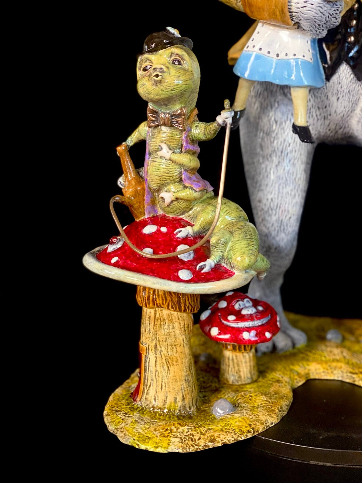 The White Rabbit figurine features a green caterpillar in a hat and bowtie lounging on red and white mushrooms, holding a hookah pipe, with a nod to Alice-inspired whimsy and partially visible girls legs in the background.