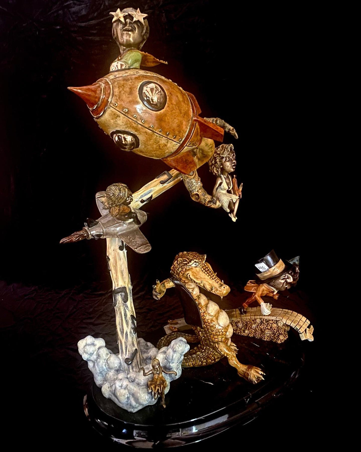 The whimsical bronze sculpture, If I Was a Sculptor, showcases a top-hatted crocodile reminiscent of Elton Johns style. Nearby, a rocket ship and miniature airplane add to the dynamic and surreal scene with playful figures evoking the spirit of Rocket Man.