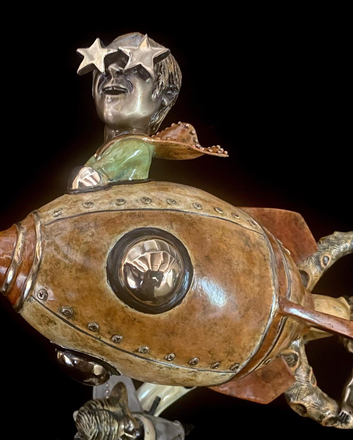 The whimsical bronze sculpture If I Was a Sculptor features a person with star-shaped glasses, reminiscent of Elton John, riding a small stylized rocket. Metallic accents and a circular window detail the craft against a black background.
