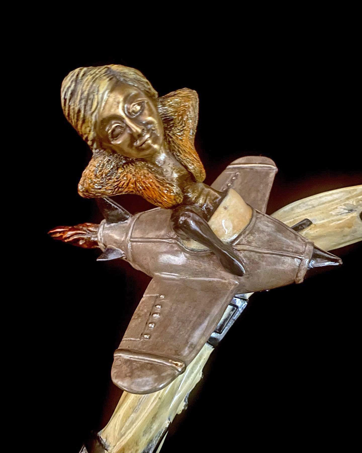 The bronze sculpture, If I Was a Sculptor, portrays a woman seated elegantly on a stylized airplane with flames bursting from its tail. Her detailed facial features and textured hair stand out against a sleek black background, reminiscent of Elton Johns Rocket Man.