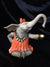 The bronze sculpture, Im a Little Teapot, features an elephant in a red dress, orange bow, and ruffled collar. Sitting on its haunches against a dark background, it gracefully balances a brass-colored cup with its trunk.