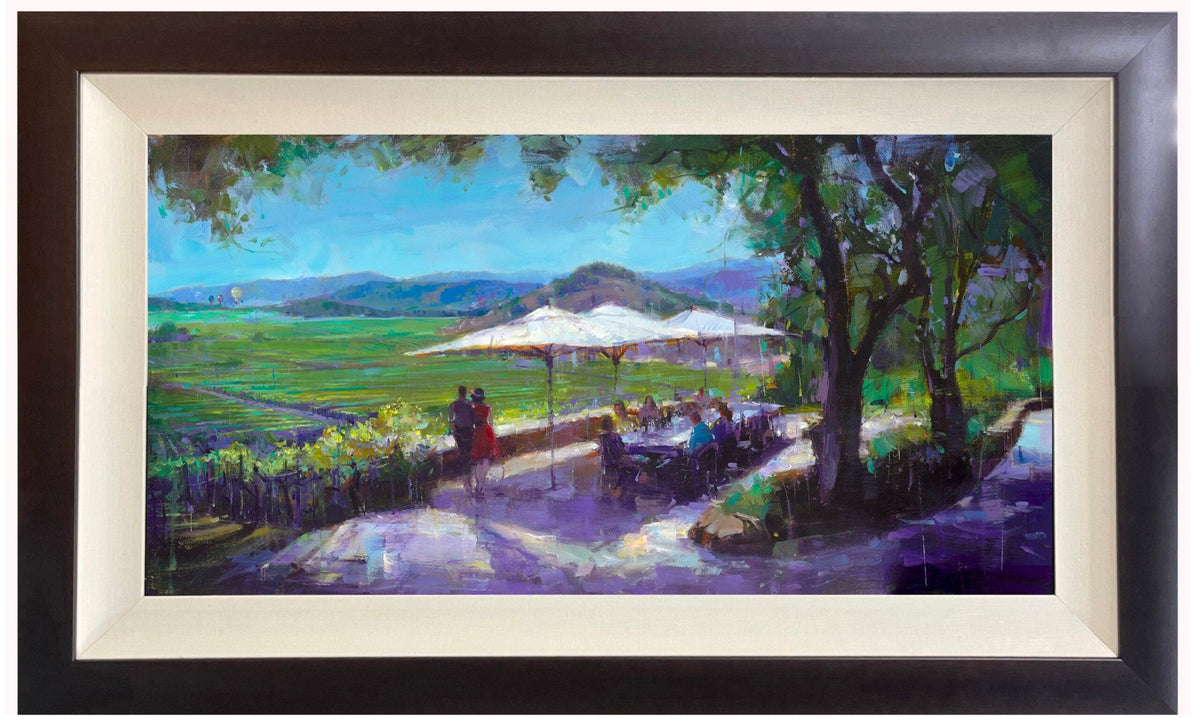 The framed painting Joie de Vivre captures a scenic outdoor dining scene in Napa Valley with people under umbrellas, lush green hills, and a clear blue sky. Vineyards stretch beneath while trees cast dappled light on the path and terrace.