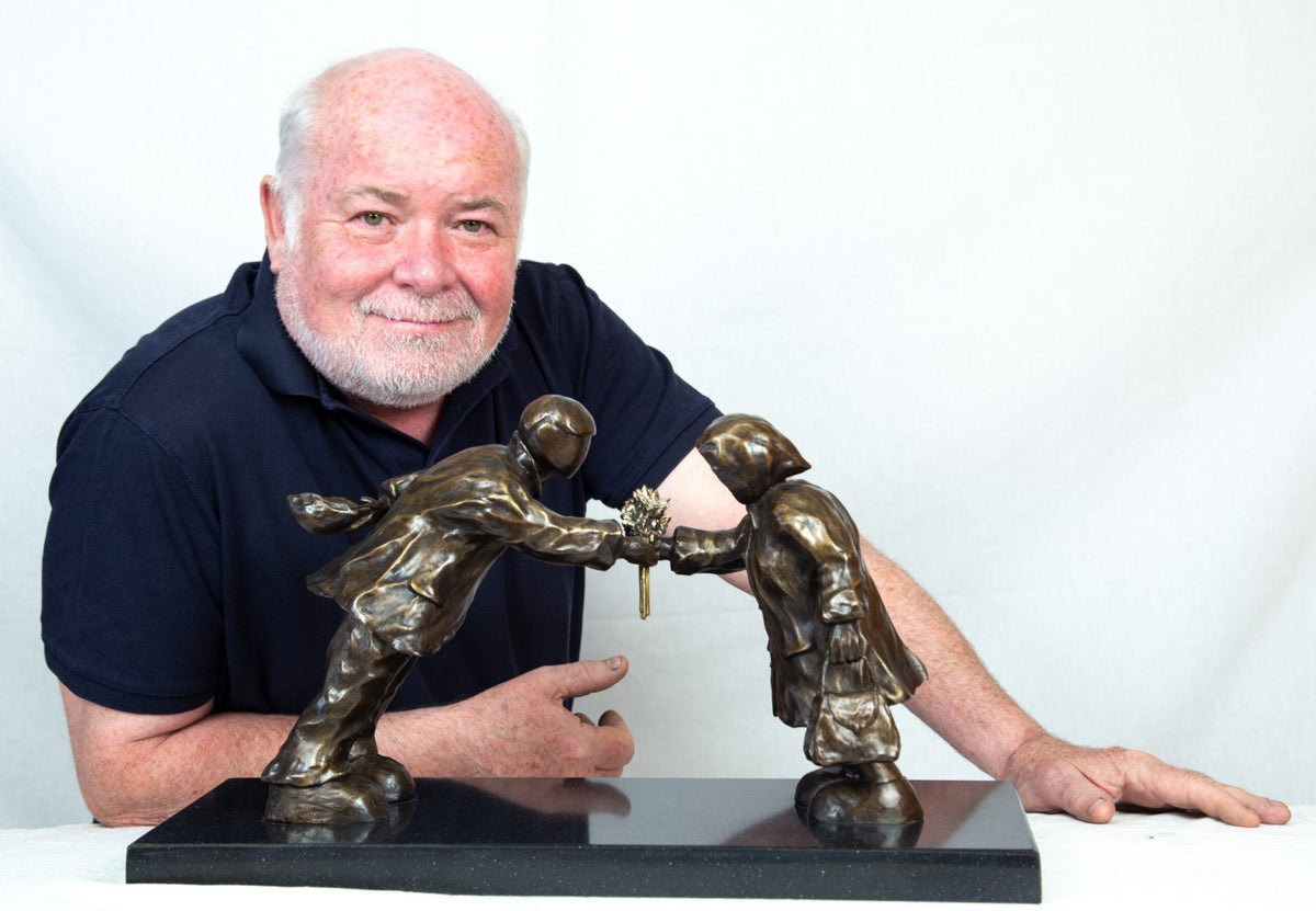 A man with a white beard in a black shirt poses with the Leaning In For Love sculpture by Mackenzie Thorpe, featuring one figure offering flowers to another, elegantly resting on a black base.