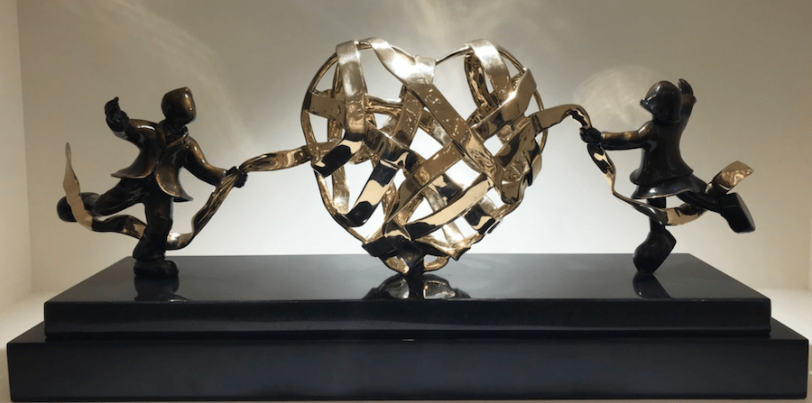 The limited edition sculpture Lovers Entwined by Mackenzie Thorpe features two stylized bronze figures holding ribbons around an intricately woven heart, all mounted on a glossy black base with soft backlighting.
