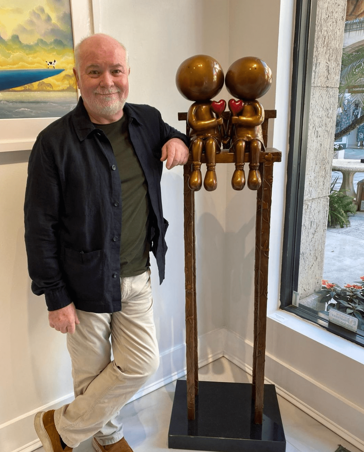 A man stands indoors next to the tall bronze Lovers on the Bench Mackenzie Thorpe sculpture—two round-headed figures with red hearts, side by side on a high bench by a window—at Marcus Ashley Gallery.