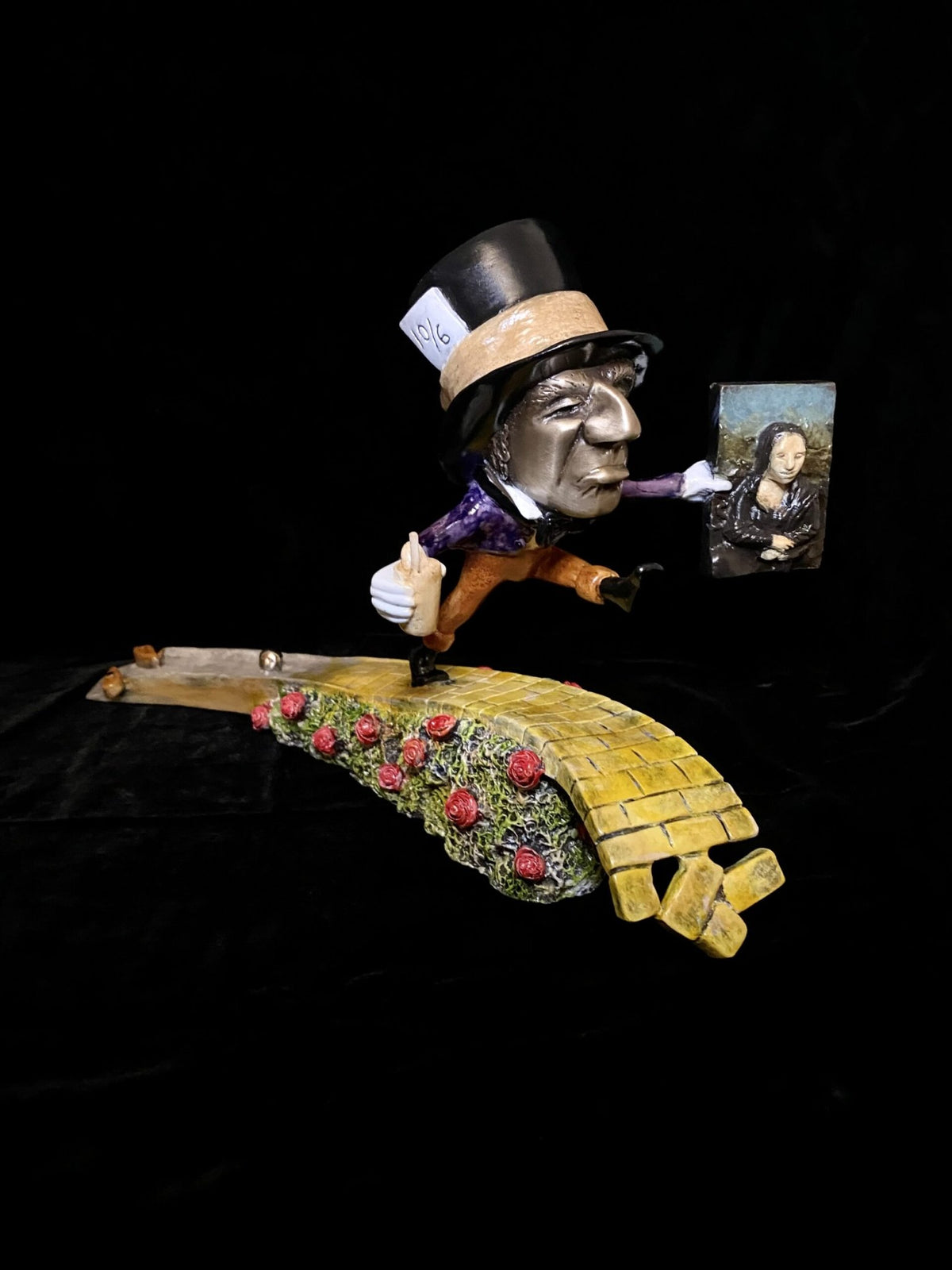 The Mad Hatters sculpture captures the whimsical essence of Lewis Carrolls character, featuring a bronze Mad Hatter with a 10/6 tag and framed Mona Lisa. It stands on a yellow brick road with vibrant red roses, evoking a surreal theme against a dark background.