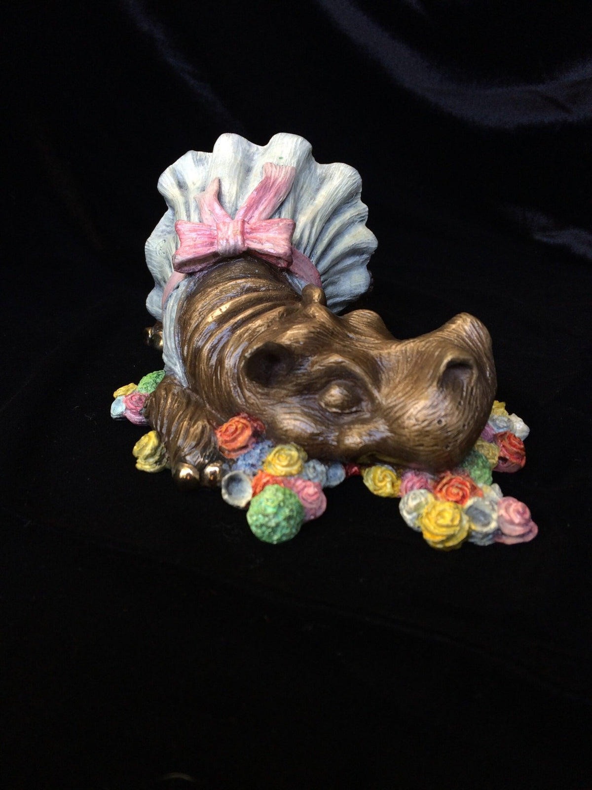 The Mary, Mary Quite Contrary bronze sculpture features a whimsical hippopotamus in a blue tutu and pink bow among vibrant flowers, all against a striking black background, capturing the playful charm of nursery rhymes.