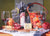A still life shows two glasses of red wine, a bottle of Members Only, blue cheese, pomegranate, knife, corkscrew, and basket of persimmons on a table—an original watercolor in Eric Christensen’s style.