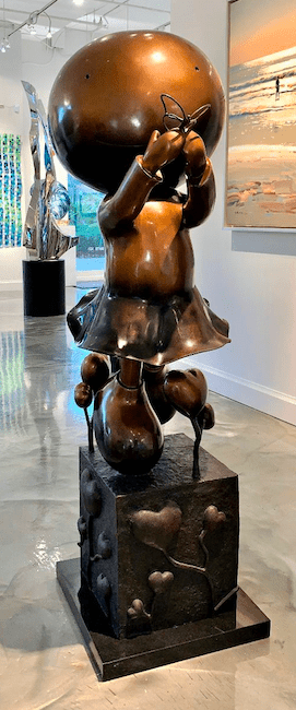 The limited edition sculpture My Butterfly-Bronze depicts a child holding a butterfly on a pedestal with heart-shaped decorations. Its displayed in a bright gallery alongside various paintings and sculptures.