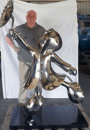 A person stands behind Mackenzie Thorpes My Sunshine-Stainless, a limited edition sculpture crafted from shiny stainless steel. The stylized figure holding a sunflower gleams on its black base, set against a partially visible white backdrop.