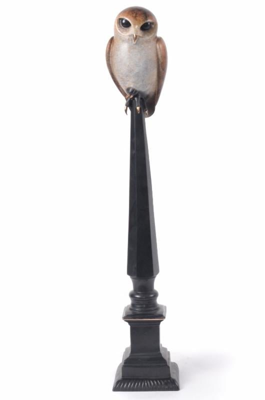 Napoleon, a realistic bronze owl statue with brown and white feathers, perches atop a tall black ornate pedestal against a white background.