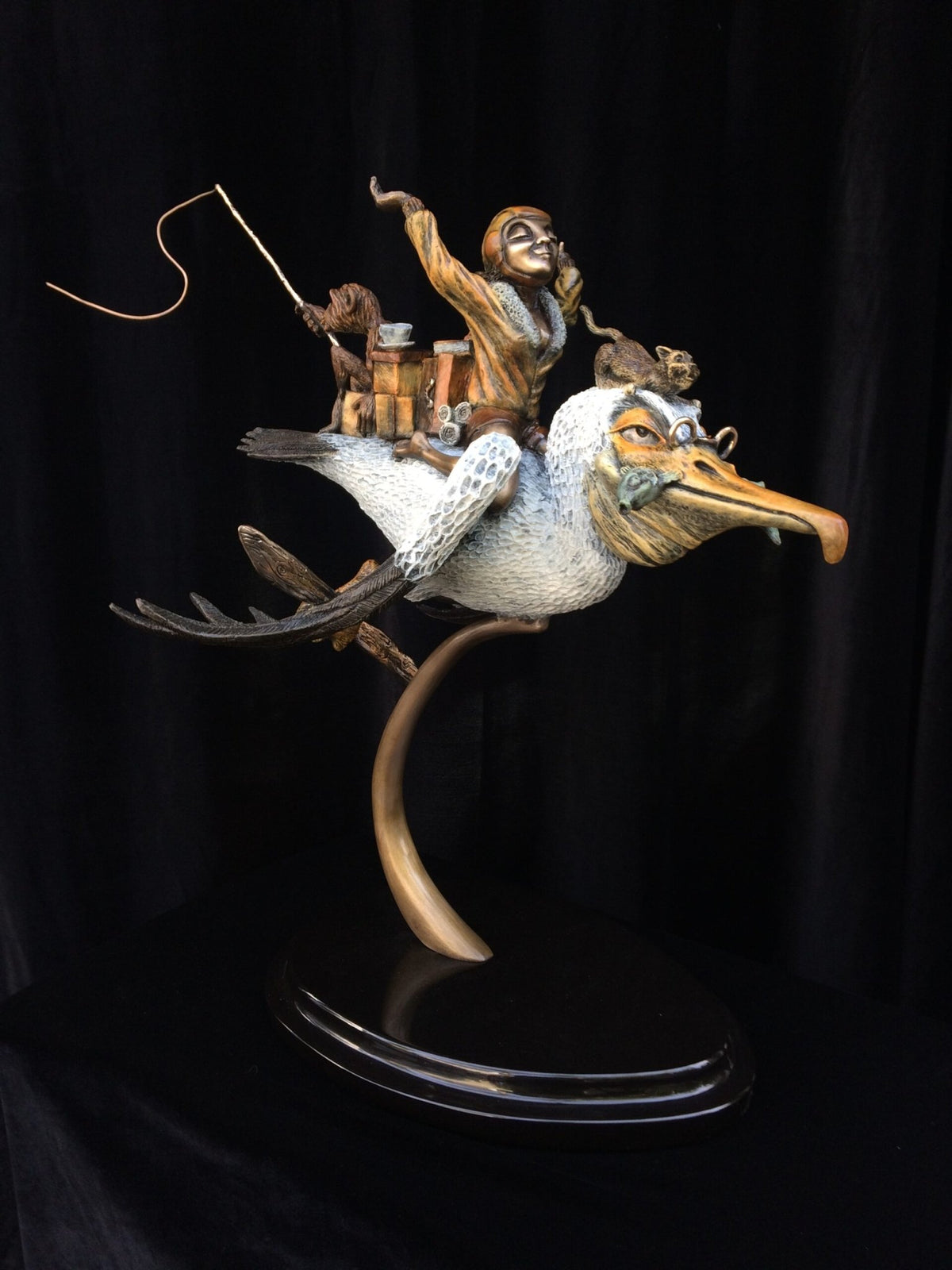 A whimsical bronze sculpture titled Ode To Amelia Earhart depicts a daring monkey riding a large bird with a long beak, followed by smaller monkeys, mounted on a stand against a dark backdrop, capturing an imaginative adventure.