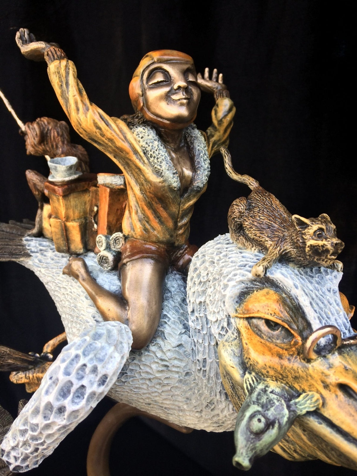 The bronze sculpture Ode To Amelia Earhart features a vivacious figure in a fur-collared jacket, arms raised, riding a fantastical creature with textured wings and a rodent on its head against a vibrant dark backdrop, capturing an adventurous spirit.