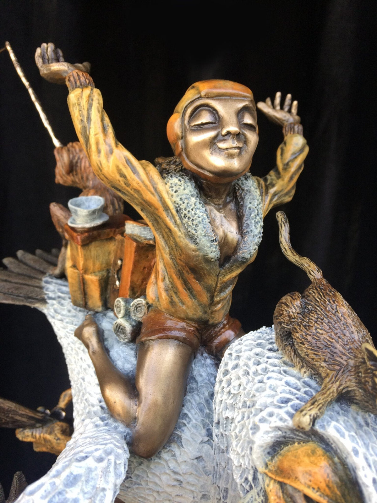 The Ode To Amelia Earhart sculpture features a joyful figure in a vintage aviator outfit on a bird with textured wings, while a top-hatted monkey and playful cat complete the whimsical scene.