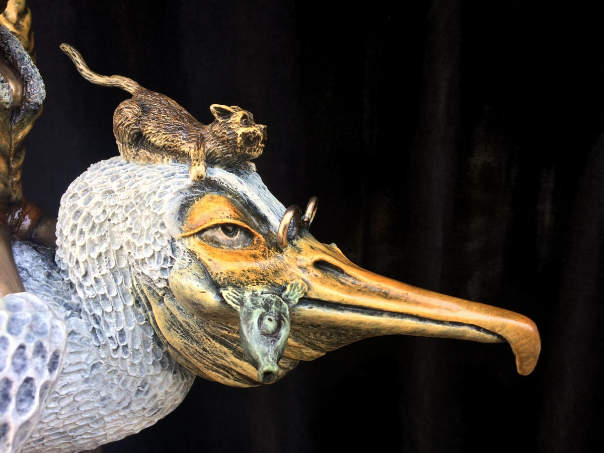 Ode To Amelia Earhart, a bronze sculpture, showcases a birds head with a long beak and textured feathers, topped by a small, cat-like animal. The black background highlights its adventurous spirit and intricate details.