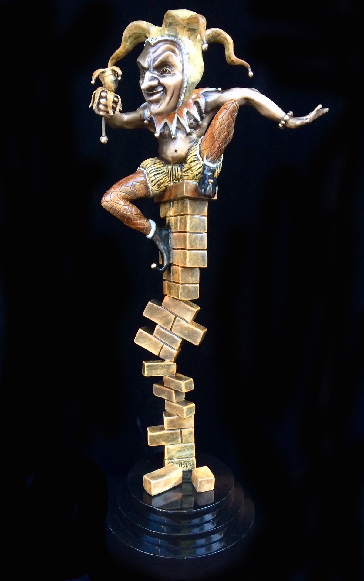 The bronze sculpture Surely, You Jest? by Paul Lotz features a whimsical jester playfully balancing on large, gold Jenga-like blocks, adorned with a vibrant hat and holding a scepter while raising one leg in delight.