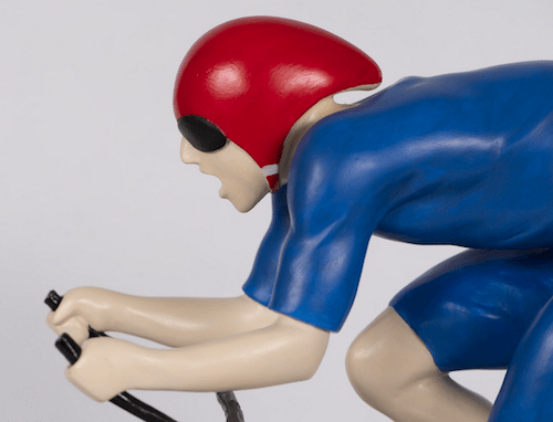 Mackenzie Thorpes limited edition bronze sculpture, The Fastest, portrays a focused cyclist in a blue outfit and red helmet with a black visor, leaning forward on black handlebars against a plain backdrop, capturing the spirit of the Tour de Yorkshire.