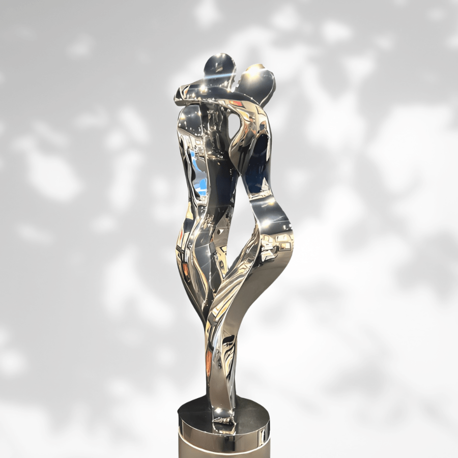 The Embrace - Polished Steel sculpture, with its intertwined figures, reflects human connection through minimalist design. Its smooth metallic surface captures light and shadows, evoking movement against a blurred, leafy background.