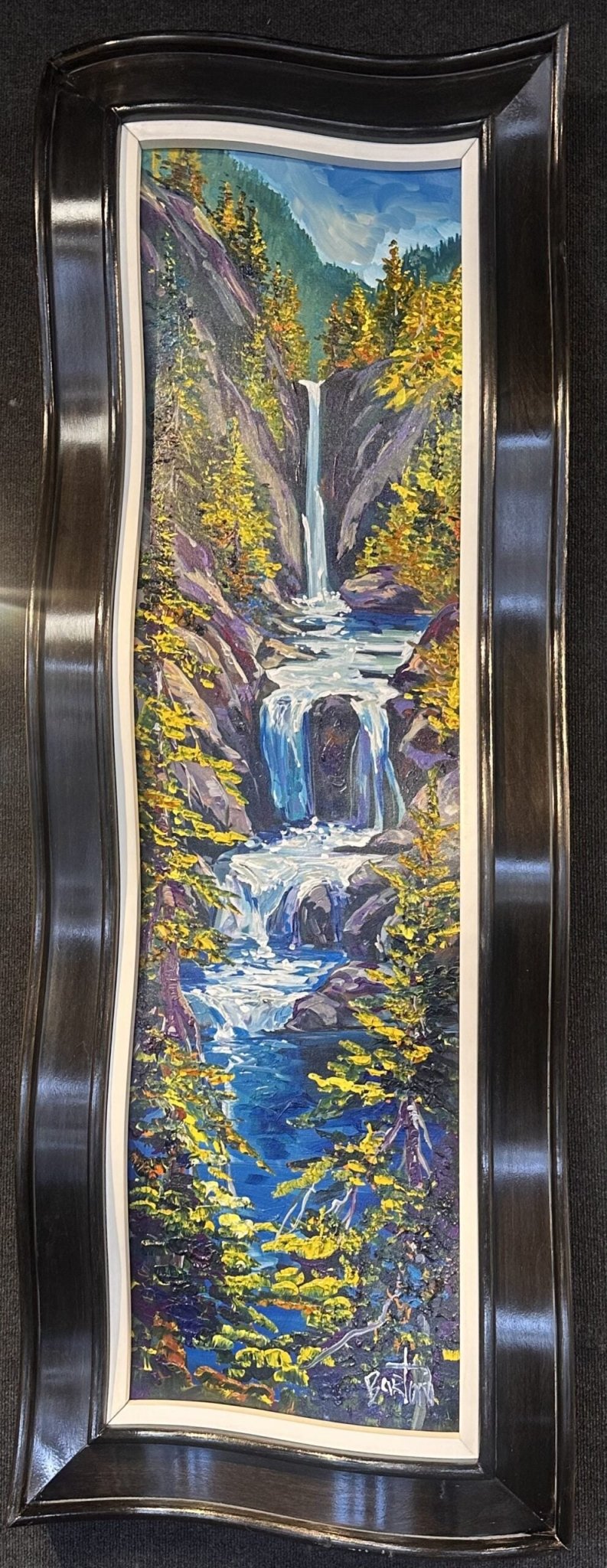 Where Life Flows is a vertical painting of a cascading waterfall through rocky terrain, embraced by lush greenery. Framed in a stylish, curvy dark frame reminiscent of Steve Bartons designs, it enhances this powerful, vibrant natural scene.