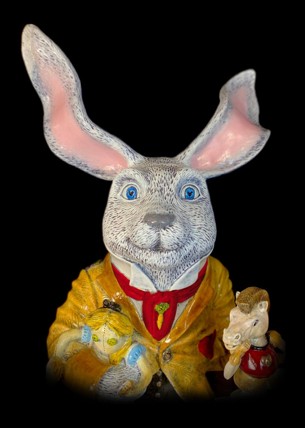 The White Rabbit is a ceramic figure featuring large pink ears, blue eyes, a yellow coat, and red tie. It holds a teddy bear and mouse and stands out against a black background, evoking Alice’s whimsical adventures.