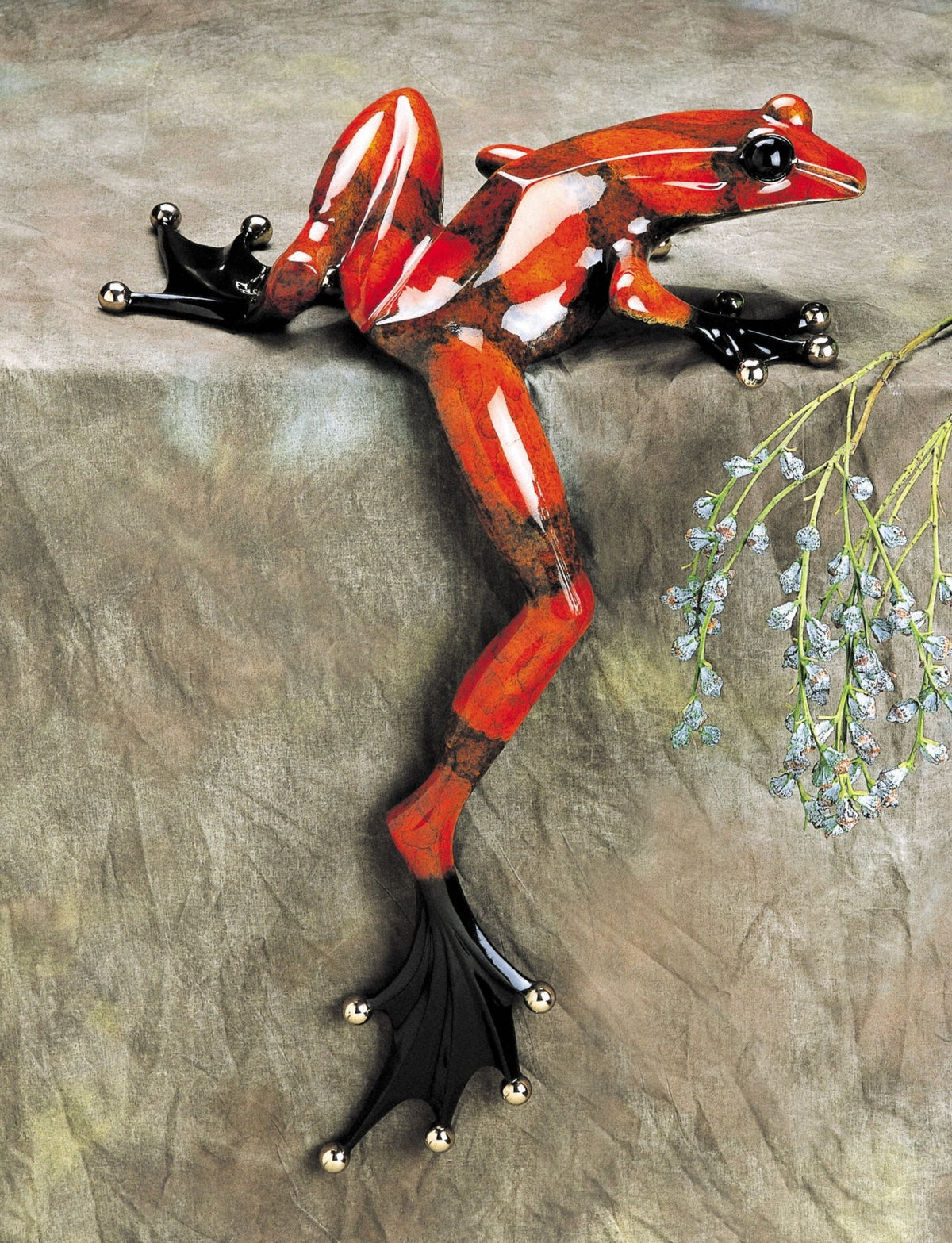 A dynamic sculpture named Willie Jump features a red and black frog with exaggerated limbs and a glossy finish, poised as if climbing. It stands against a textured gray background with nearby delicate flowers, echoing its movement style.