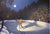 Beneath the full moon, a figure in a yellow jacket elegantly skates on the frozen pond—now a Woodland Arena. The scene evokes Rob Gonsalves art as an eager crowd watches from snowy banks, surrounded by towering trees.