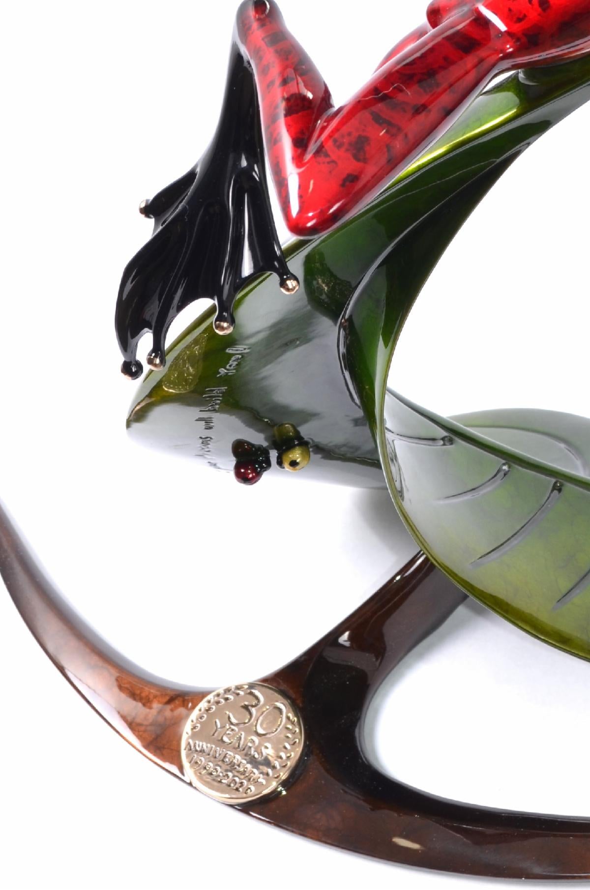 Zenith by Frogman Bronze is a vibrant red frog sculpture with glossy black and red legs, perched on a green leaf-shaped base. The brown platform displays a silver medallion commemorating the 30th anniversary.