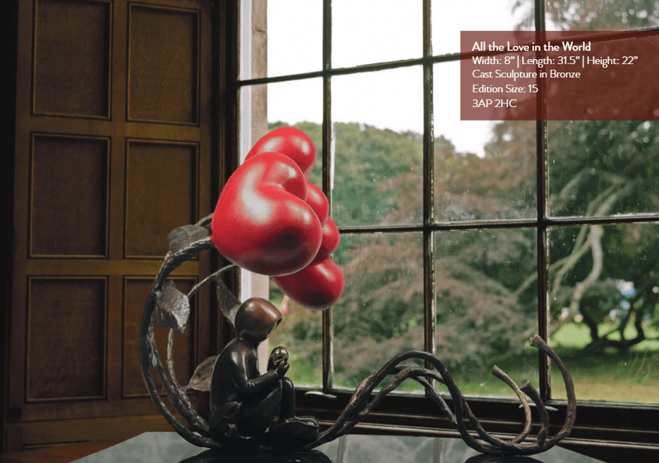 The bronze sculpture All The Love In The World, featuring a seated figure with red heart-shaped balloons, is showcased indoors by a window at Marcus Ashley Gallery in the Lake Tahoe art district, with details on its dimensions and edition.